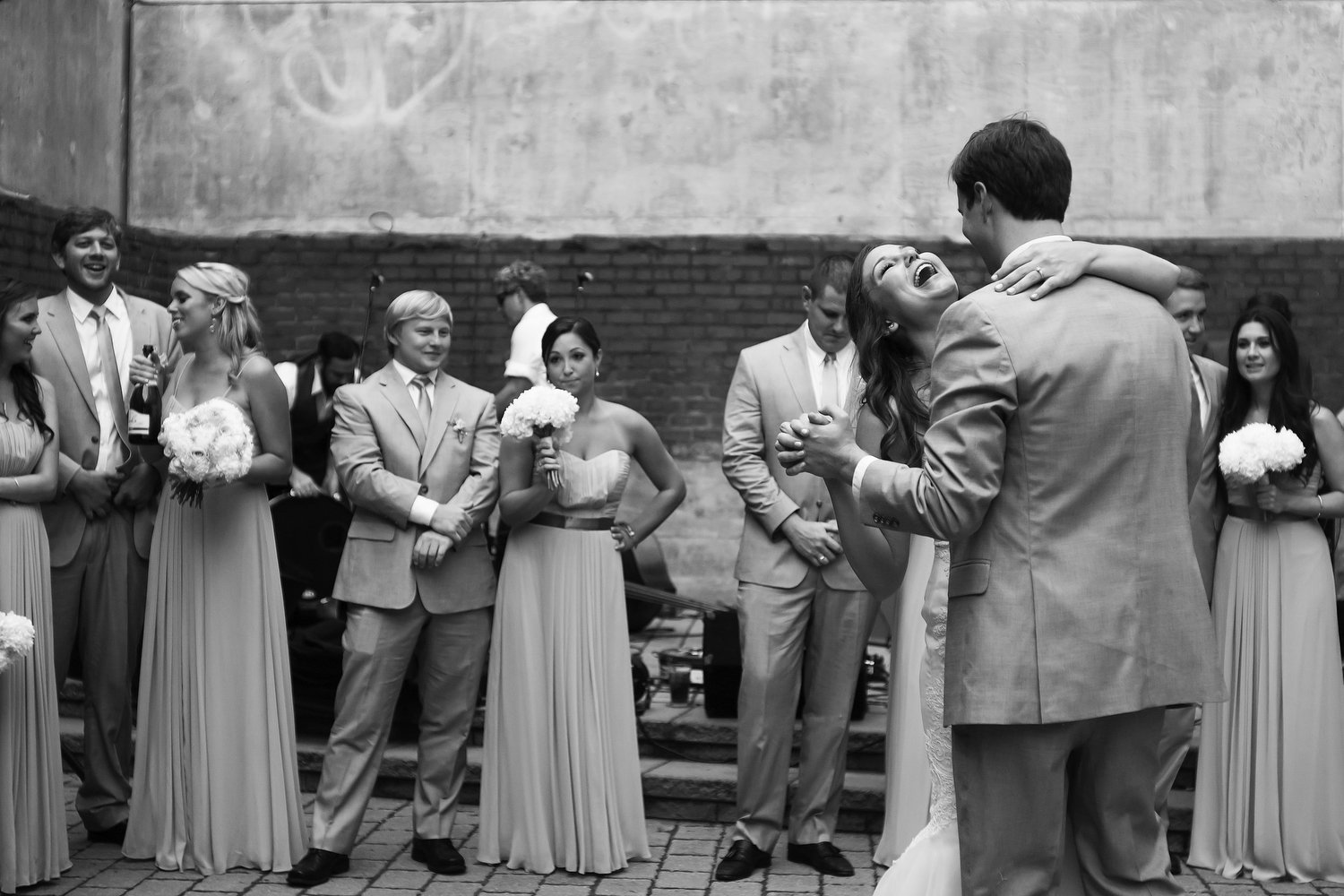 A wedding reception with a bride and groom dancing surrounded by wedding party and guests, black and white photo. She has her head tipped back laughing and her arms around his neck.