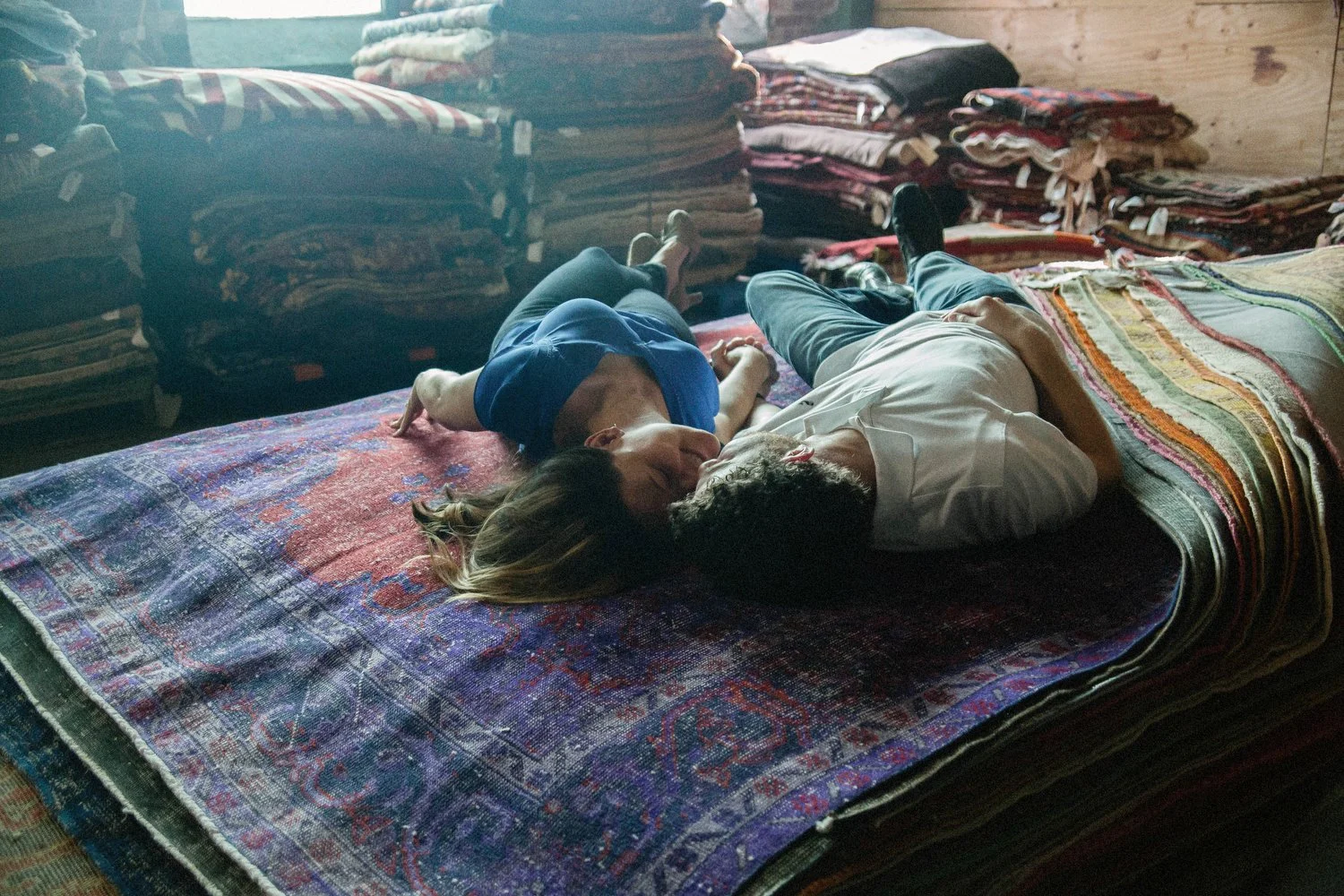 A couple is seen lying down on a stack of decorative rugs in a warehouse or place with several stacks of textiles lining the walls behind them. They are facing each other and kissing. She has on a blue short-sleeved shirt and jeans. He has on jeans a