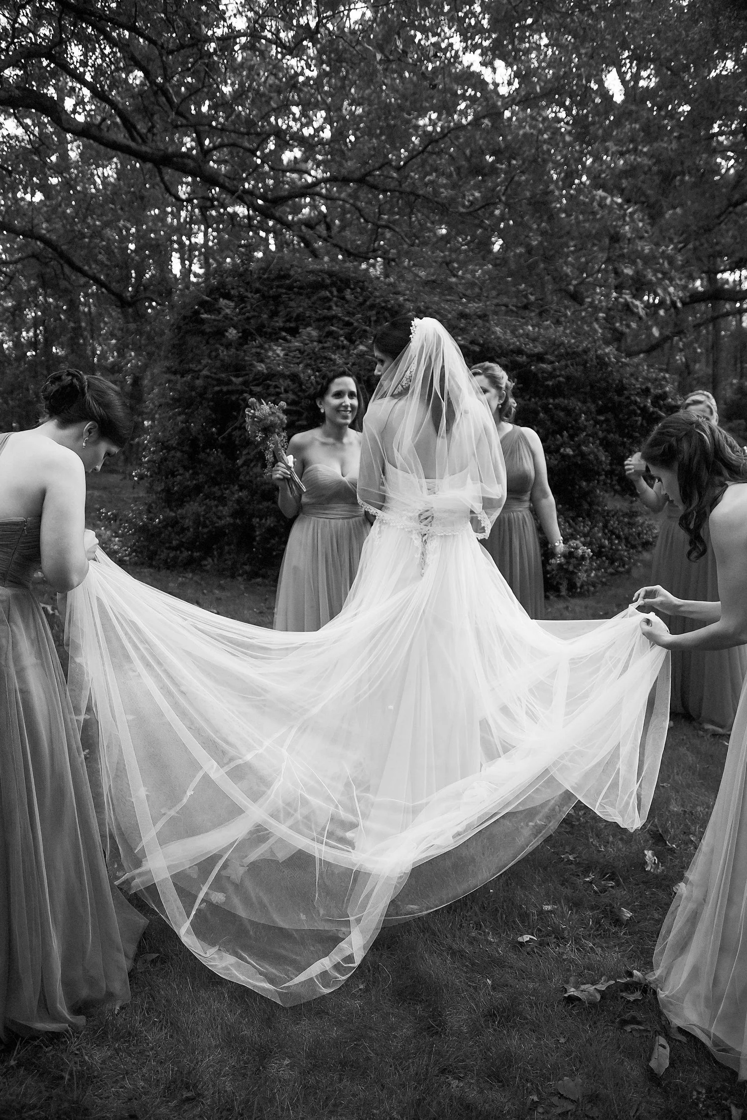 Black and white photo of a bride in a wedding dress and veil surrounded by bridesmaids holding her train outdoors in a garden.