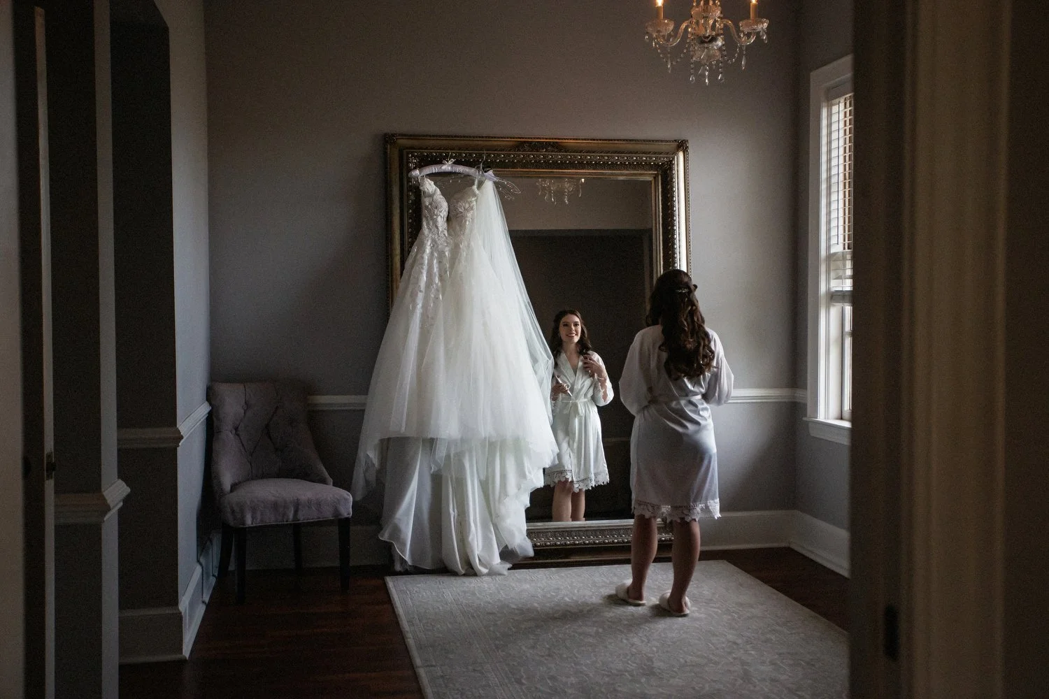 A woman in a white satin robe looks at her reflection in a large mirror, with a wedding dress hanging above her. The room has gray walls, a chandelier, a window, and a chair.