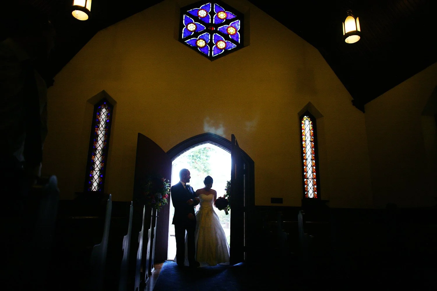 Silhouette of a bride and her father standing at church doorway, backlit by outside light, with stained glass windows and interior lighting visible. A large geometric hexagon window is illuminated by blue and yellow stained glass on the wall above th