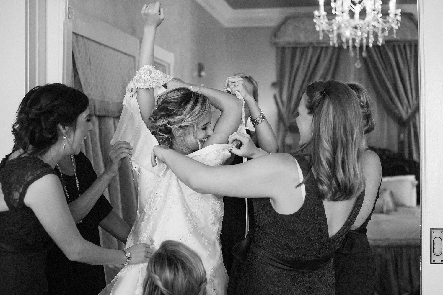 A bride is surrounded by women helping her put on her wedding dress in a room with elegant curtains and a chandelier. She's looking to her right with her arms up over her head as they pull her dress down around her.