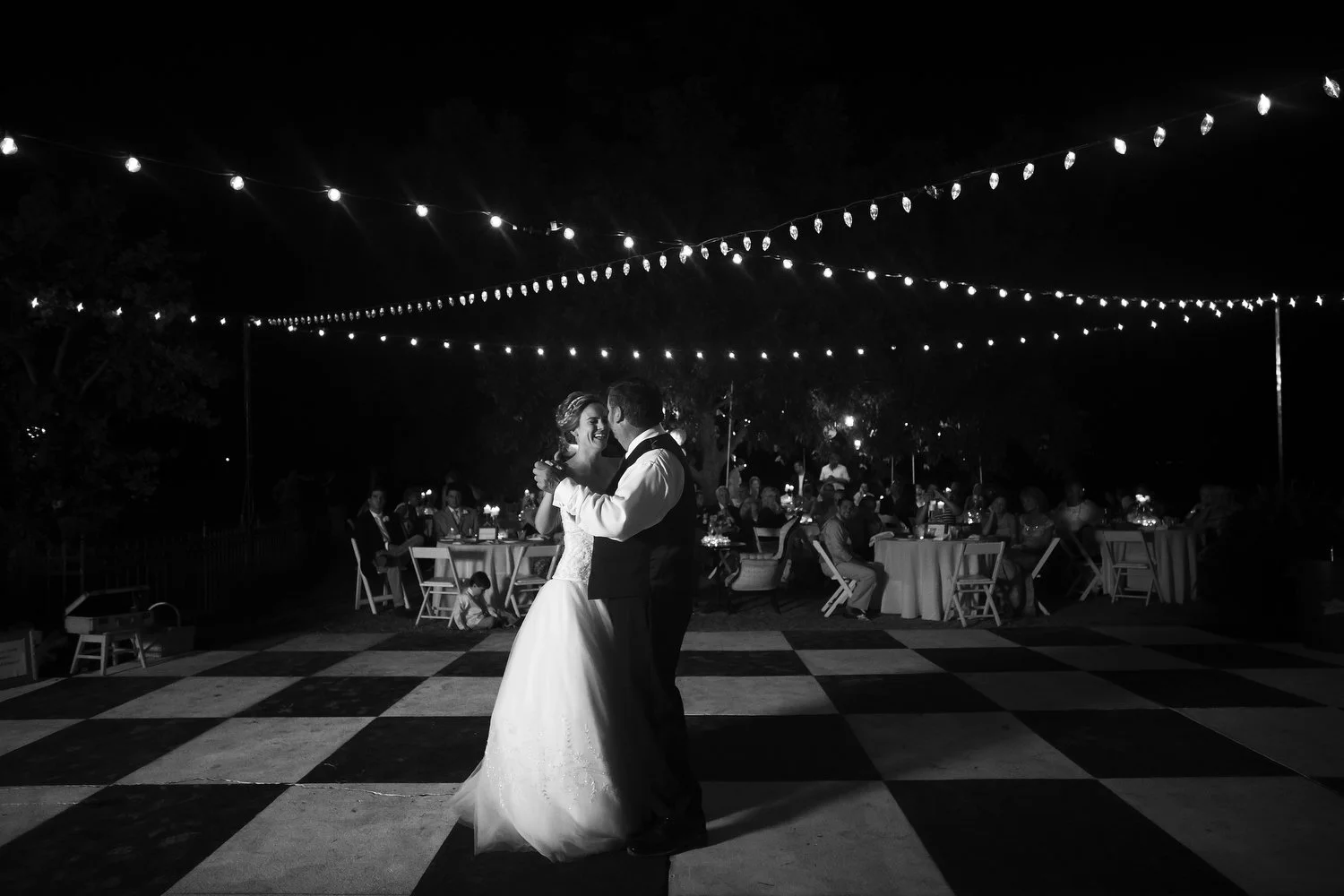 Black and white image of a couple on a black-and-white checkerboard dance floor. The dance floor is outside and strands of twinkle lights criss-cross above them. The bride has on a white wedding dress and the man, a little older so perhaps her father