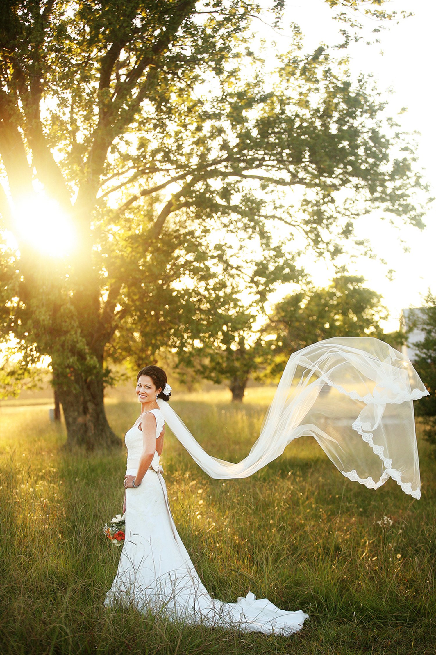 A smiling bride in a white wedding dress standing in a grassy field with trees and the sun setting behind her. Her long lacy veil blows off to the side of her.