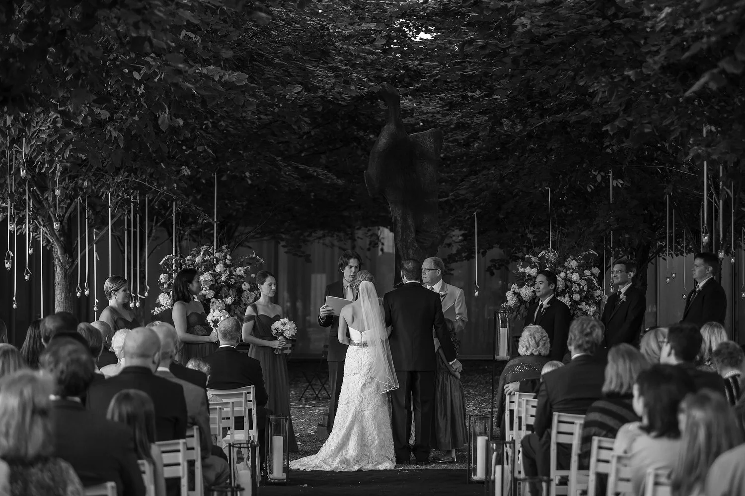 A black-and-white photograph of a wedding ceremony taking place outdoors under trees, with the bride and groom standing at the altar, surrounded by officiants, bridesmaids, groomsmen, and guests seated in chairs. The view is taken from the back of th
