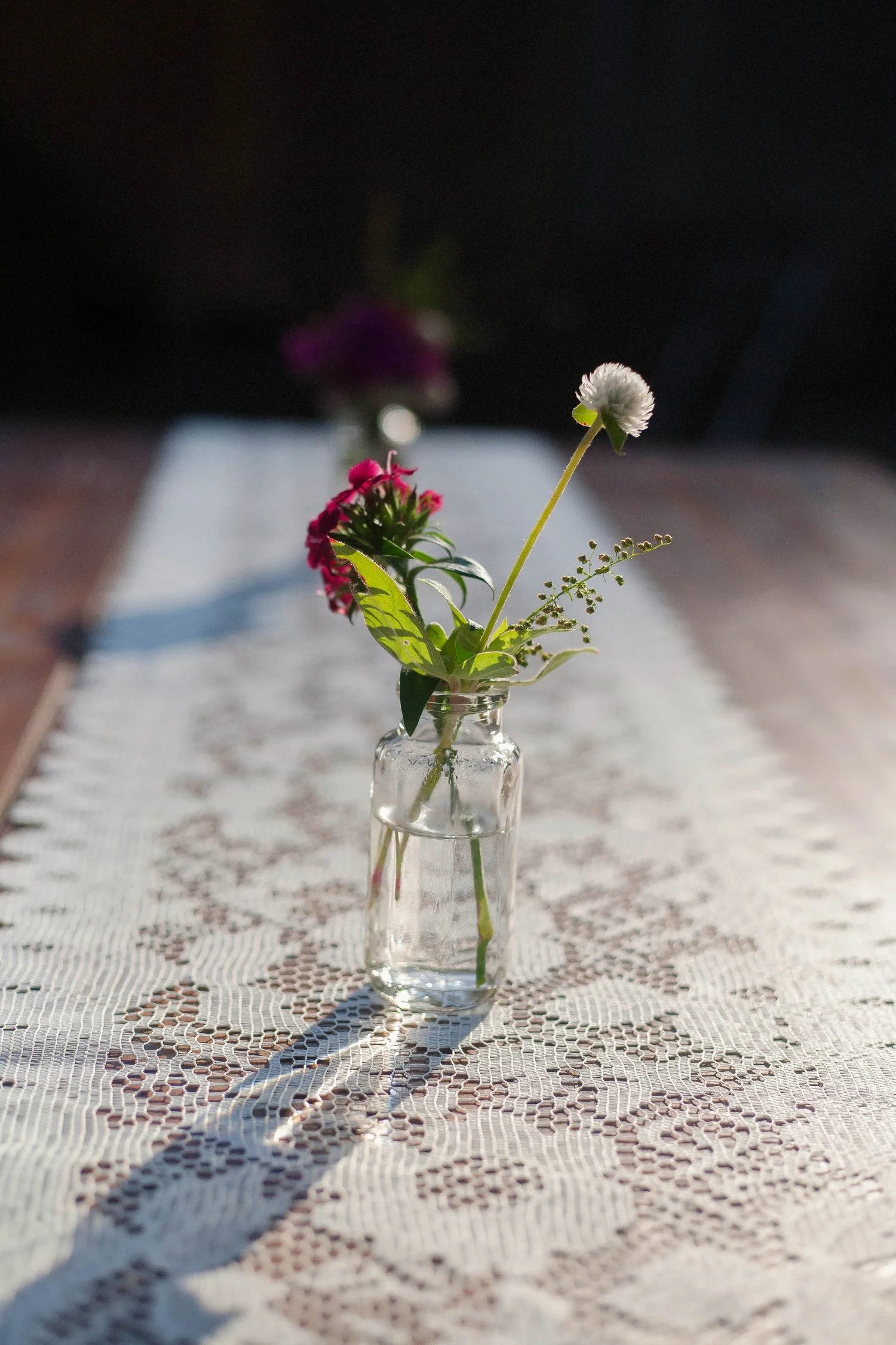 Small white and purple flowers are in a small clear jar of water on a lace runner on an outdoor table. The flowers and jar are being hit by late-day sunlight.