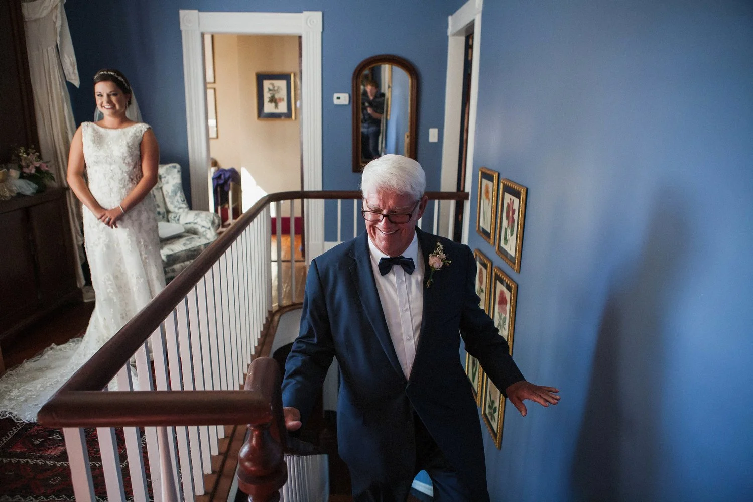 A woman in a white wedding dress standing near a staircase, smiling, as an older man in a tuxedo with a boutonniere walks up the staircase to see his daughter in her dress. The walls are painted blue and decorated with framed floral prints.