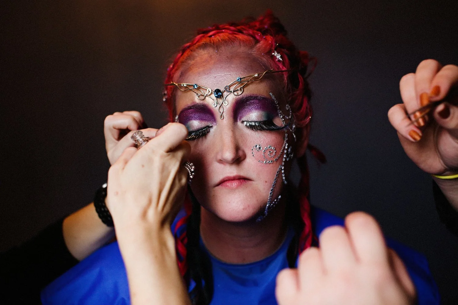 Woman with purple eyeshadow getting false eyelashes applied, wearing decorative face jewelry, and a blue top, in a dark setting.