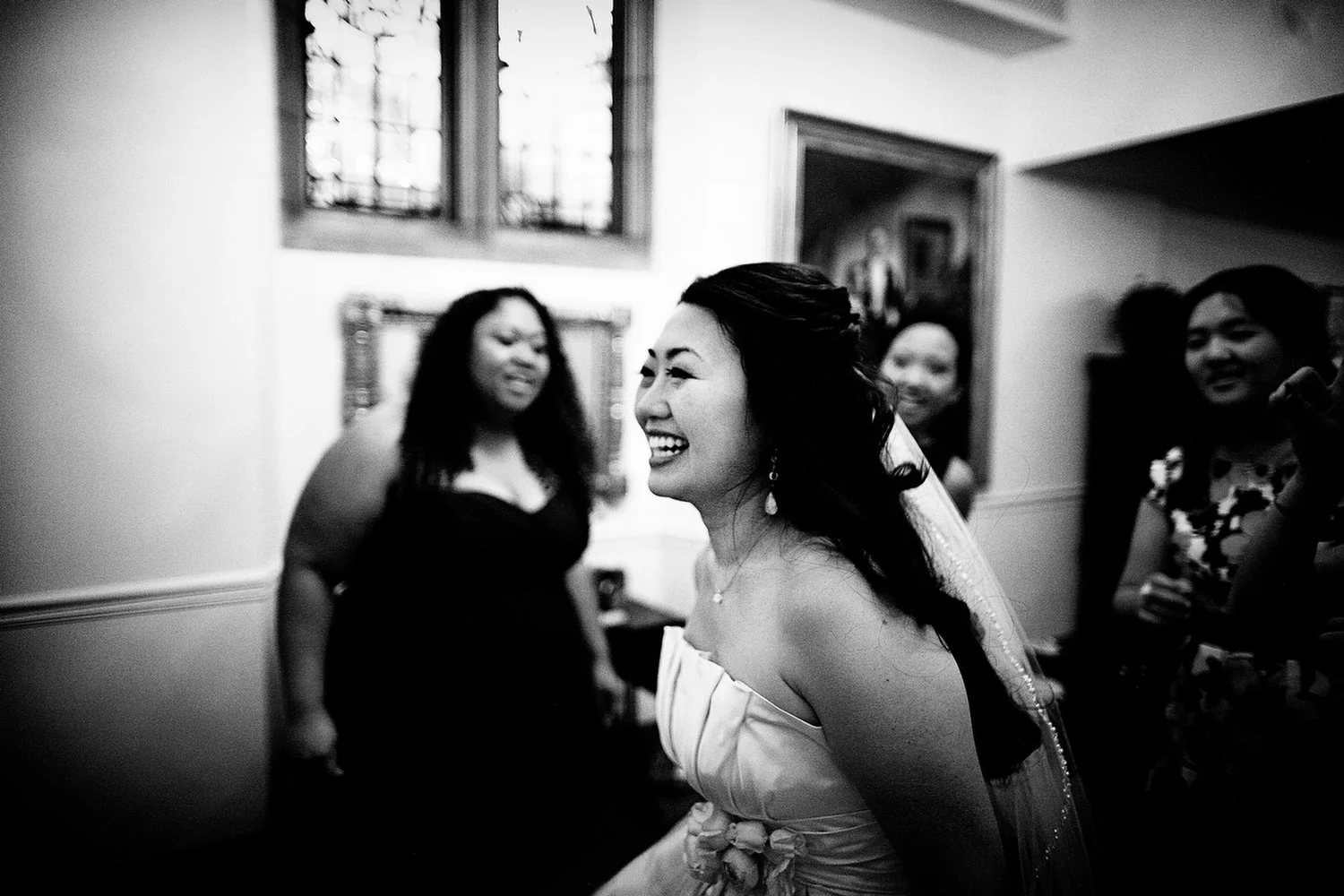 Black and white photo of a joyful bride with long dark hair, wearing a strapless dress with a floral belt and earrings, smiling and surrounded by women in a room with stained glass windows.