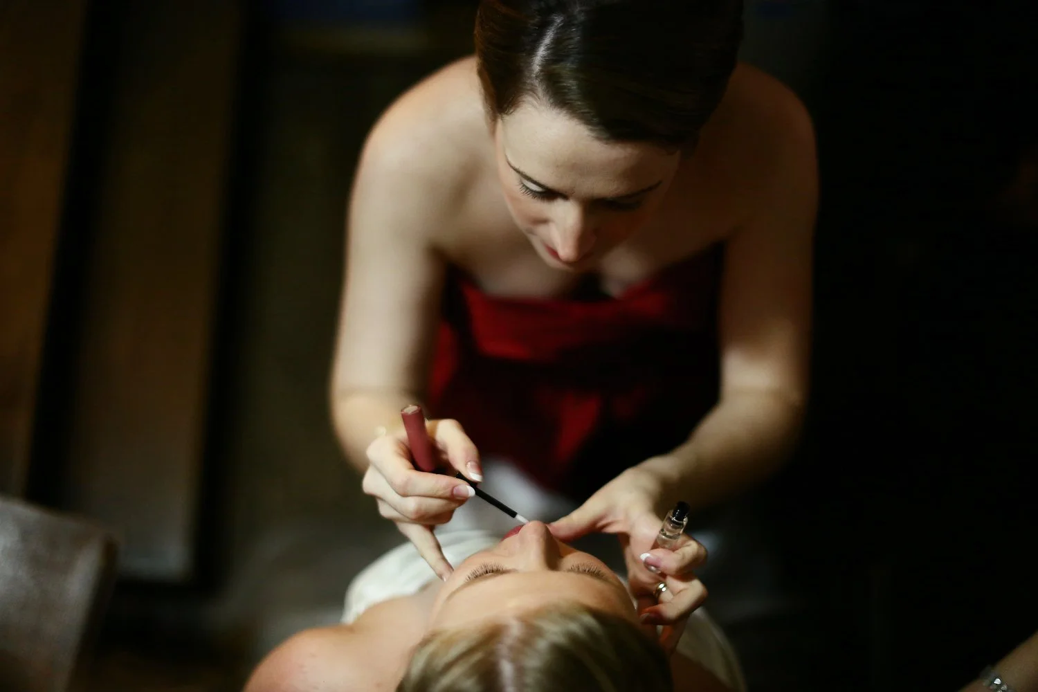 A woman in a red dress applying dark pink lip gloss on a bride wearing white. The image is taken from above and looking down at the women's eyes.