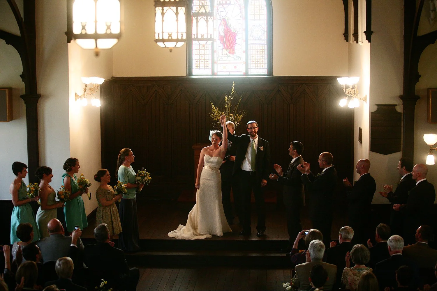 A wedding ceremony taking place in a church with the bride and groom at the center, the bride raising her hand while the groom holds her hand. Bridesmaids and groomsmen stand on either side, clapping and holding bouquets. The congregation in the pews