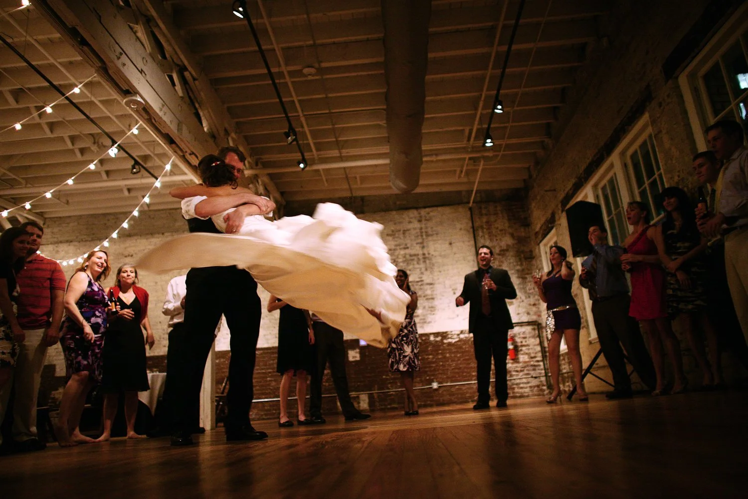 A man in black dress pants and a white dress shirt swings a woman in a long white dress around him on the dance floor in a room lit by twinkle lights. There are industrial pipes above and old brick walls behind them. Her dress is flying around her as