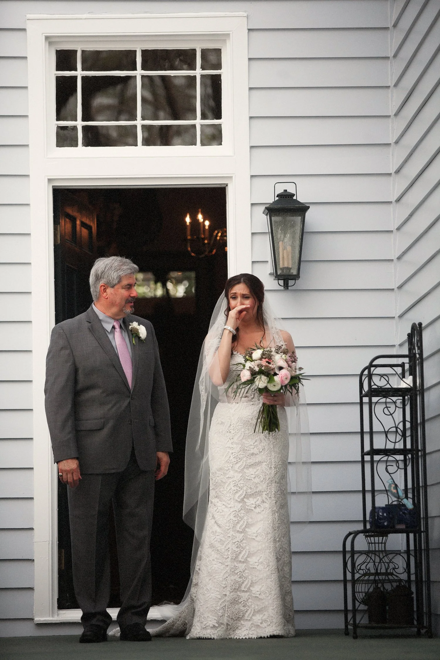 A bride in a lace wedding gown holding a bouquet of pink and white flowers, standing beside a man in a gray suit with a pink tie, outside a house with light gray siding, a wall-mounted lantern, and a window.