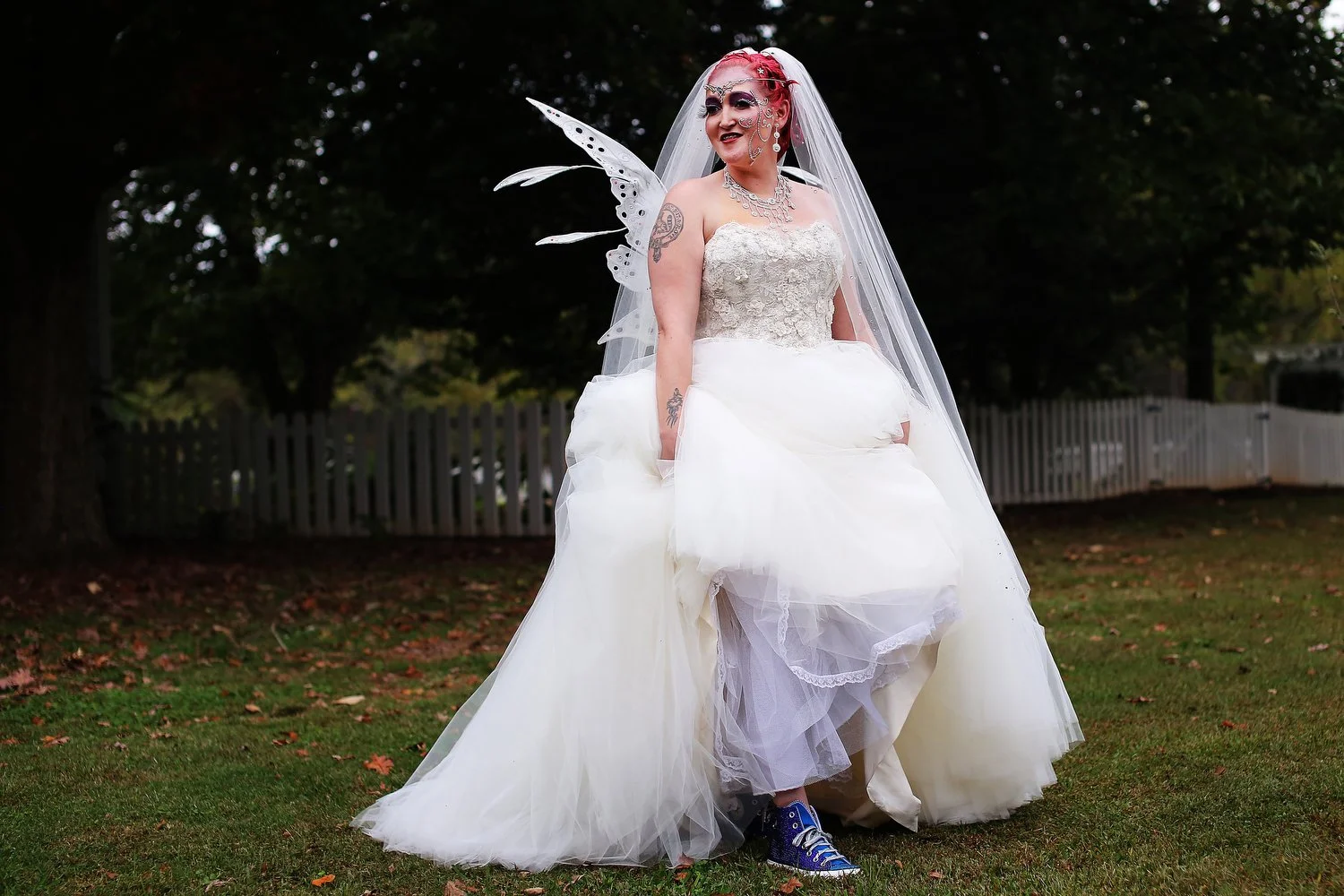 A bride with fairy wings and tattoos on her arms stands outdoors in a garden with a white picket fence in the background, wearing blue sneakers under a white wedding gown with a veil and jewels on her face.