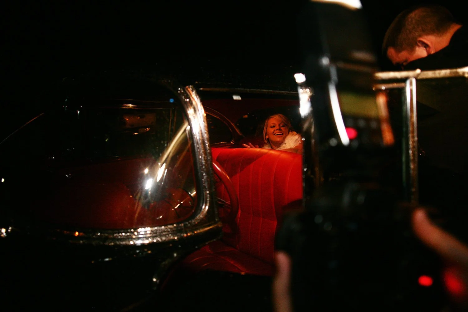 A bride in white is seen sitting in the backseat of a vintage black car with a red leather interior. The doors are open and it is dark outside. 