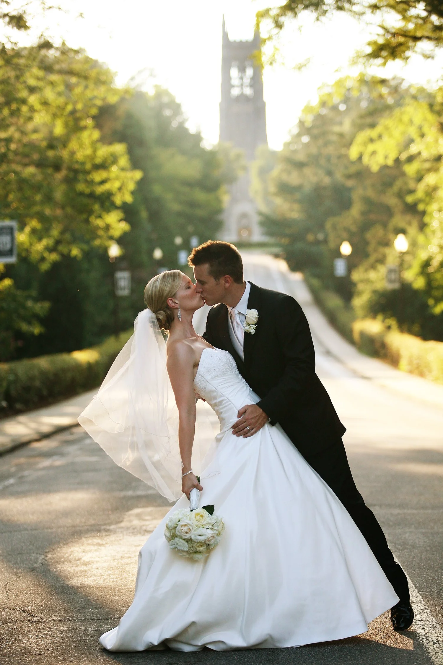 A couple stands in the foreground of an image with Duke Chapel seen in the distance down the tree-lined road. The groom is dressed in a black tux and he dips his bride slightly backward and holds her back and waist as he kisses her. She is wearing a 
