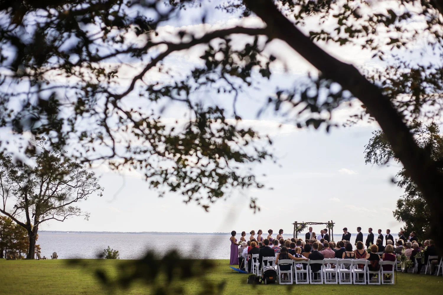 Outdoor wedding ceremony by water on the North Carolina coast with guests seated in white chairs, a wooden arch, and trees in the foreground. The photo is taken from a distance, and the ceremony is small in the frame.