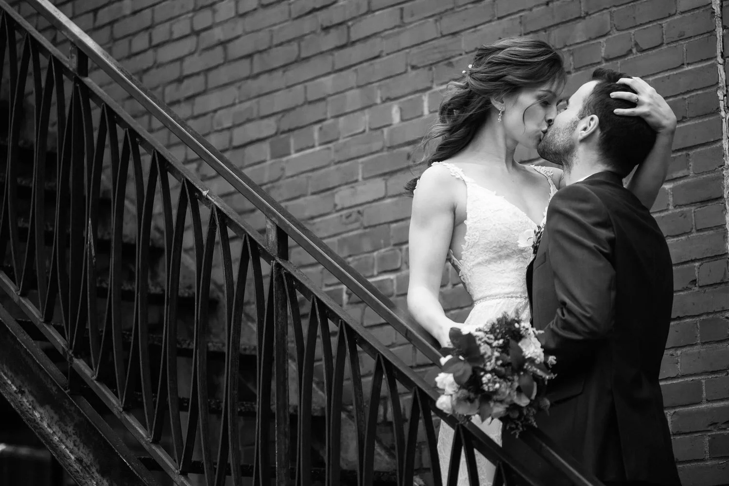 A black-and-white photo of a bride and groom kissing on a staircase in front of a brick wall. The bride is holding a bouquet and has her hand on the groom's head, while the groom is wearing a suit.