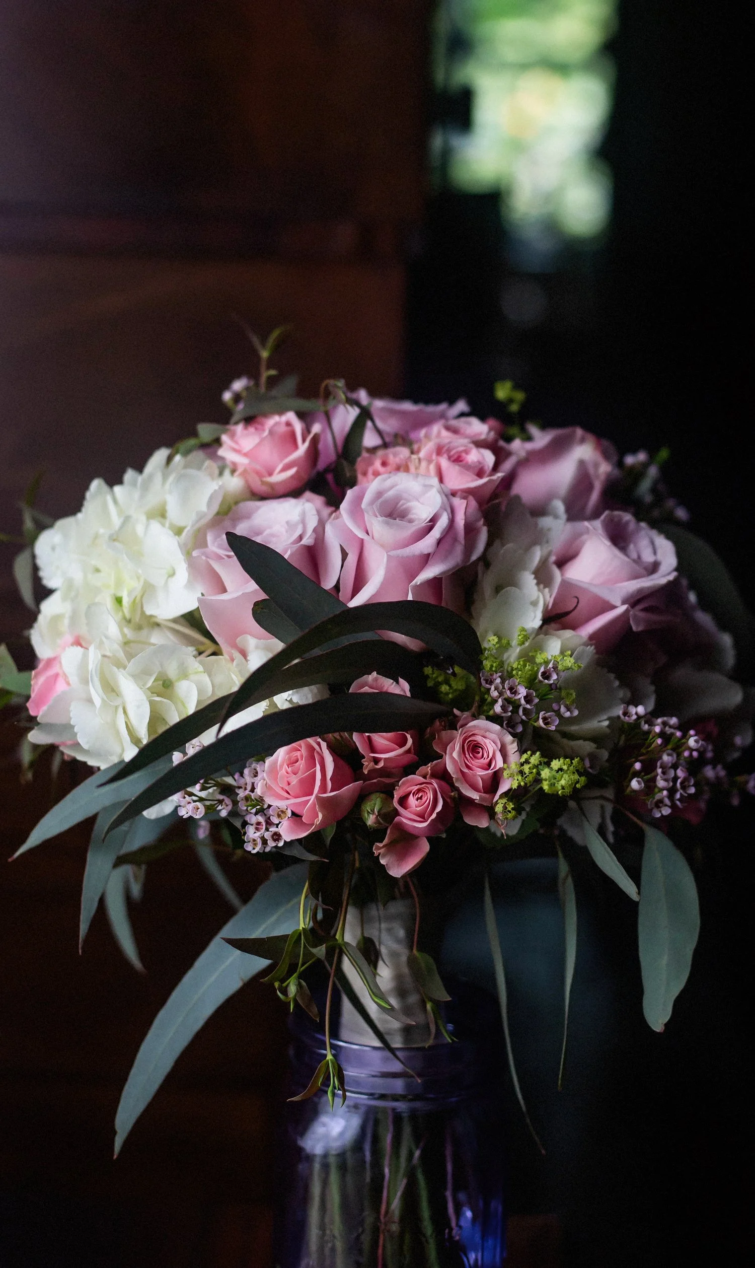 Tight vertical image of a bouquet of flowers in a glass jar. The flowers include purple and pink roses with other white floral accents. They are lit by window light.
