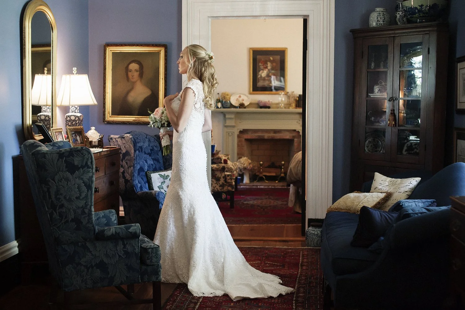 A bride in a vintage white gown stops in the hallway to look at her reflection in a gold-framed mirror on the blue wall. There are pictures on the wall behind her, a blue couch with pillows, a wooden dresser and cabinet. You can see a room behind her