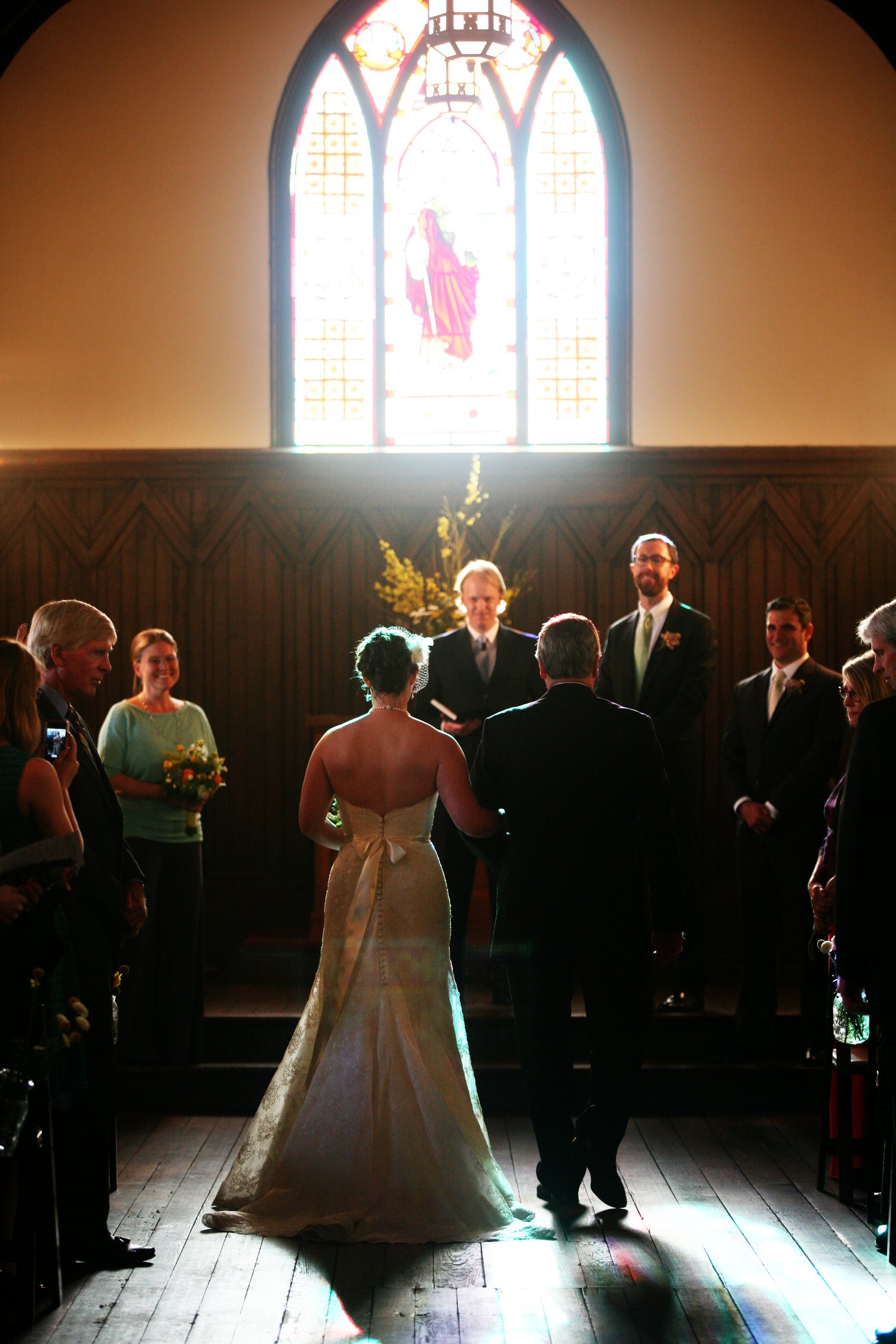 A bride and groom are standing at the altar during their wedding ceremony inside a church, with guests on each side watching, illuminated by natural light coming through a stained glass window.