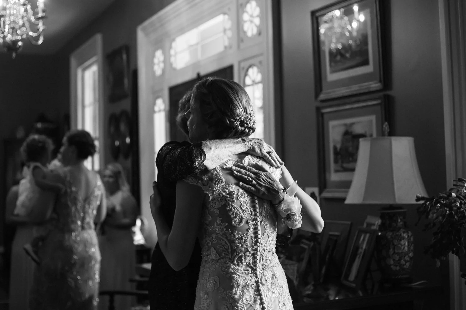 Elderly woman hugging a bride in a dimly lit room with framed pictures on the wall and a lamp on a table. The contrast of the wrinkled arms around the vintage lacy dress with dramatic button detail is evident in this black and white photo.
