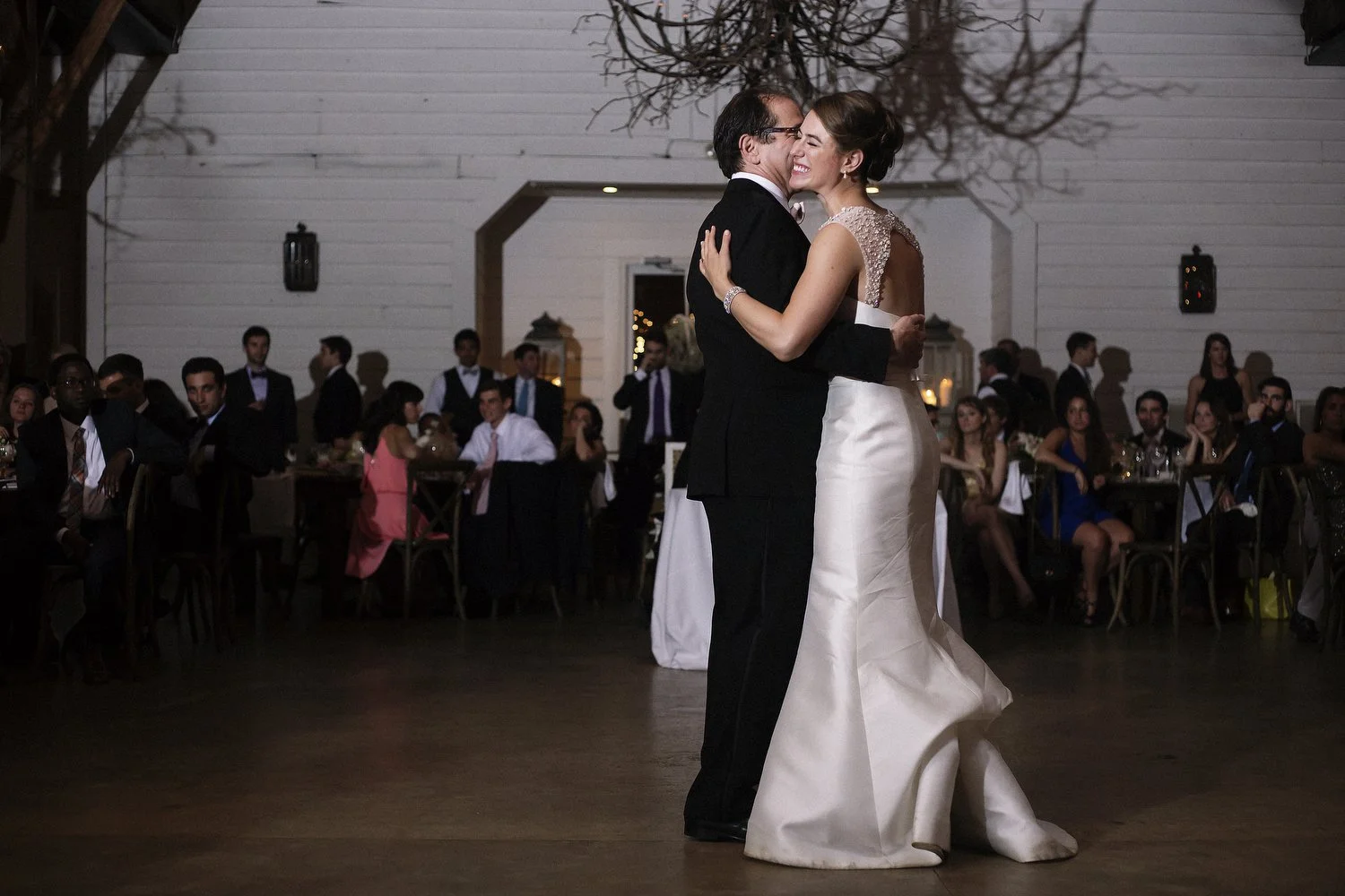 A bride in an elegant silky white wedding dress with jeweled straps clutches the back of an older man, maybe her father, in a black suit. They are on the dance floor in a dimly-lit wedding reception with several guests in the distance looking at them