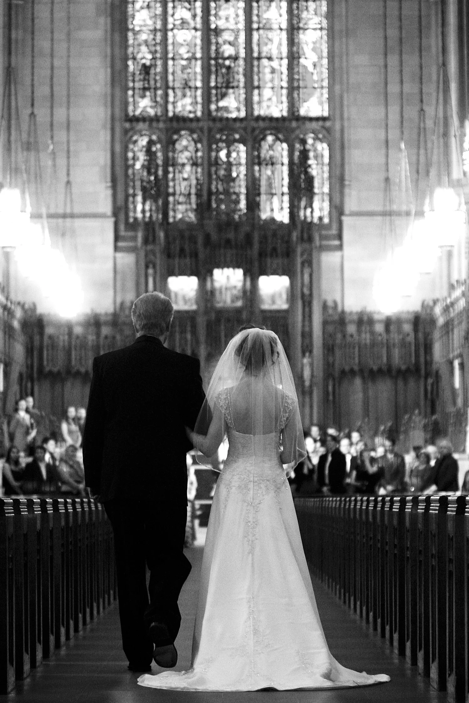 Black and white photo of a bride walking down the aisle in a church with an older man, likely her father, holding her arm during her wedding ceremony. Decorative lights line both sides of the long aisle and there are huge stained glass windows.