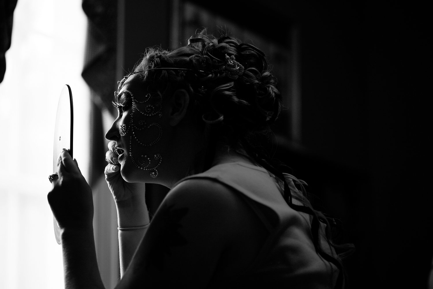A woman with elaborate jewelry and hairstyle looking into a mirror, backlit, in black and white.