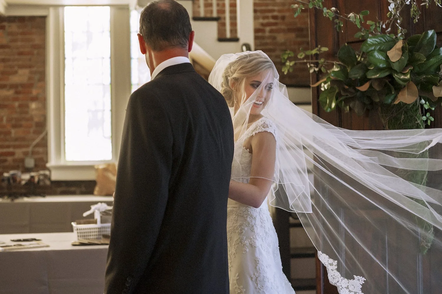 Bride in a lace wedding dress and veil smiling as she looks behind her, standing with her dad in a black tuxedo, inside a room with brick walls and large windows.