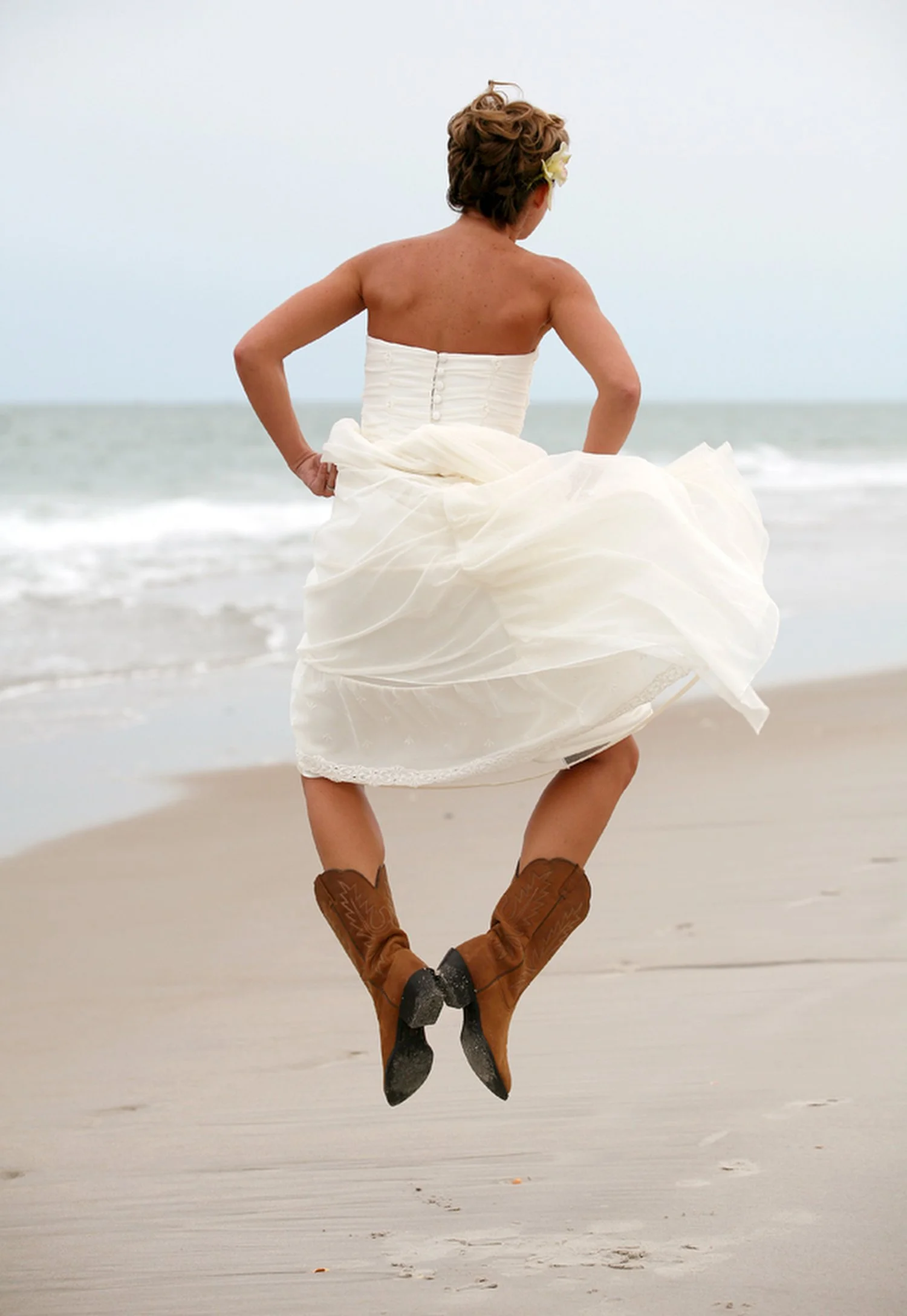 A bride is seen from behind jumping up from the sand on the beach with the ocean to her left. She is clicking her cowboy boots together at the heels and her arms are holding her dress at her waist. She has a strapless white dress and her tan back and