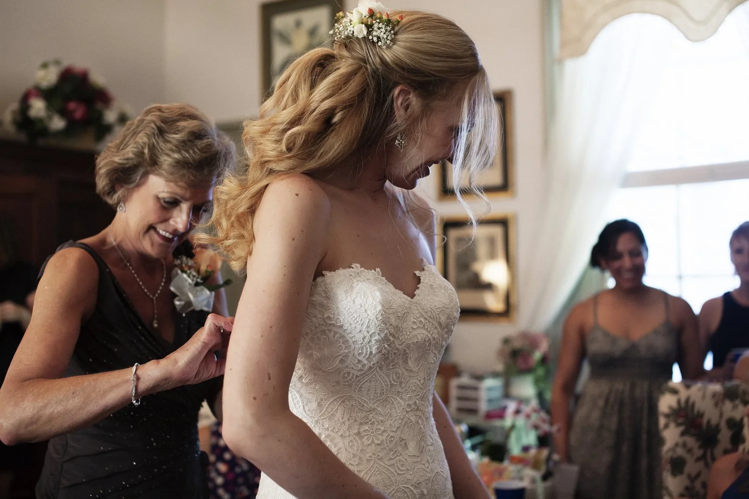 A bride in a white lace wedding dress standing indoors with a woman helping her with her dress. Other women are in the background, smiling. The room is decorated with flowers and framed pictures.