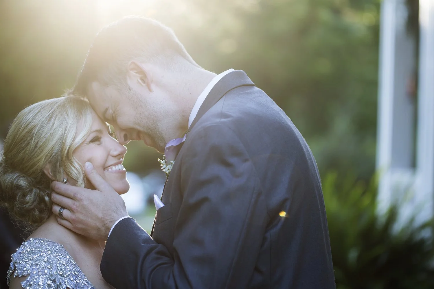 A bride and groom share a tender moment following their wedding ceremony. He has a grey suit and a purple flower on his lapel as he touches her forehead with his and places his hands on her face. She has on a vintage jeweled dress and her blond hair 
