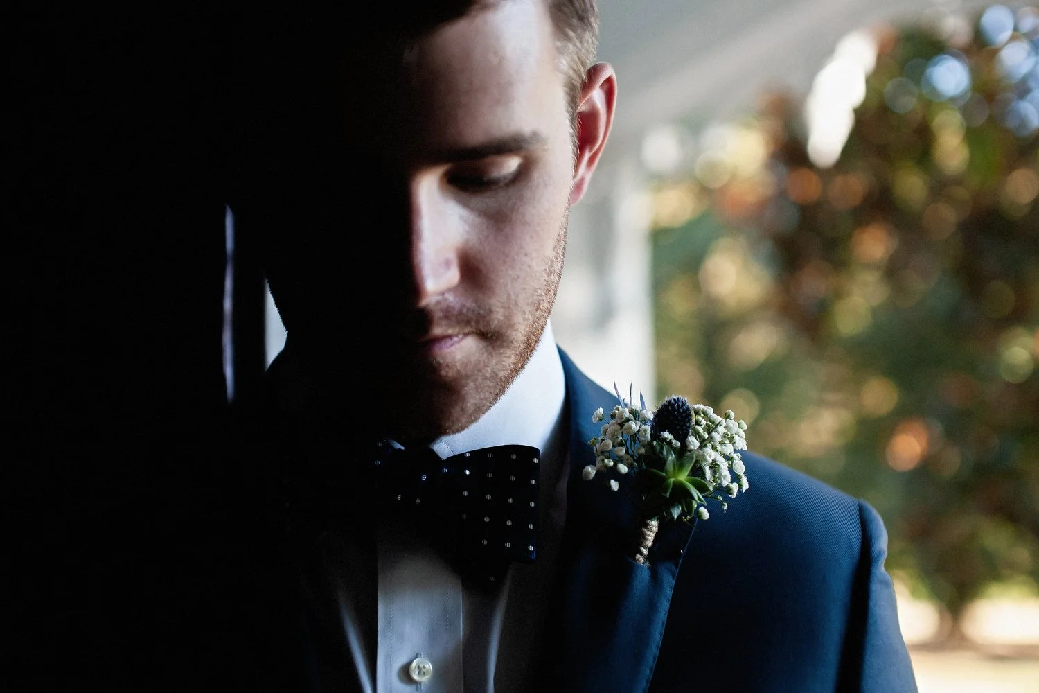 Close up image of a groom standing in a doorway with the light coming in from the outside. He looks down with a serious expression. He has on a dark blue suit jacket, white dress shirt and polka-dotted bowtie. His boutonniere has baby's-breath on it.