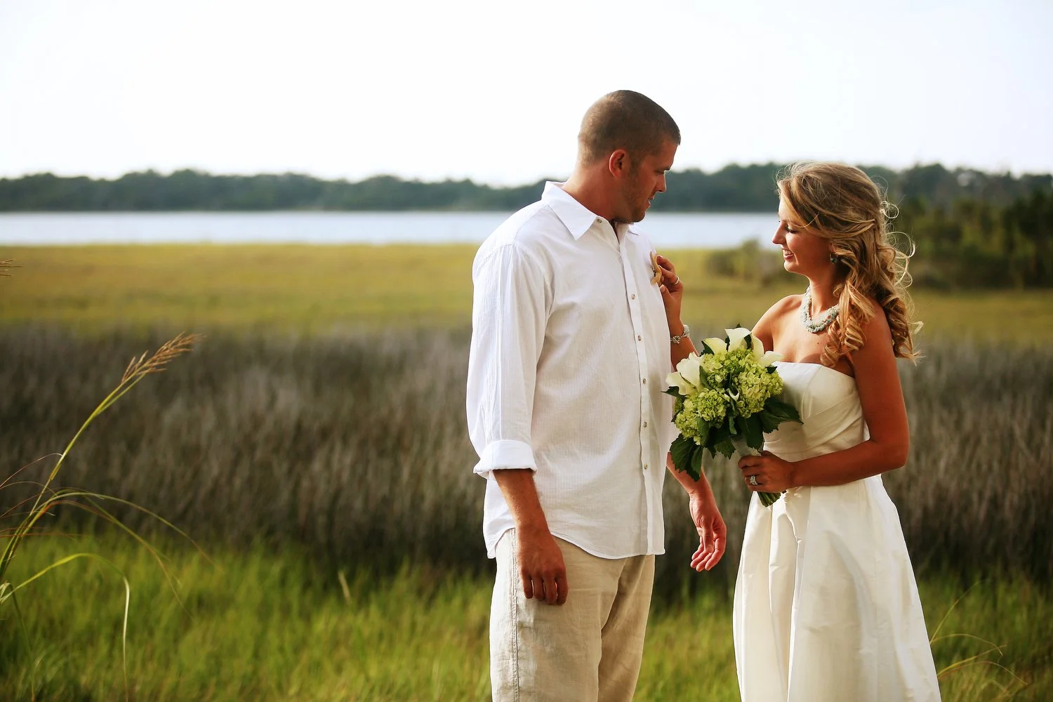 A bride and groom outdoors in a coastal setting. She is in a white strapless dress, holding a bouquet of white flowers. He is in khakis and a white dress shirt. They are in the grass with tall marsh grasses and water behind them. 