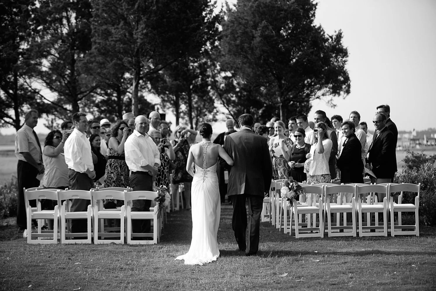 Black and white photo of a wedding ceremony outdoors, with a bride and her father walking down the aisle, surrounded by seated guests and standing people on both sides, with trees in the background.