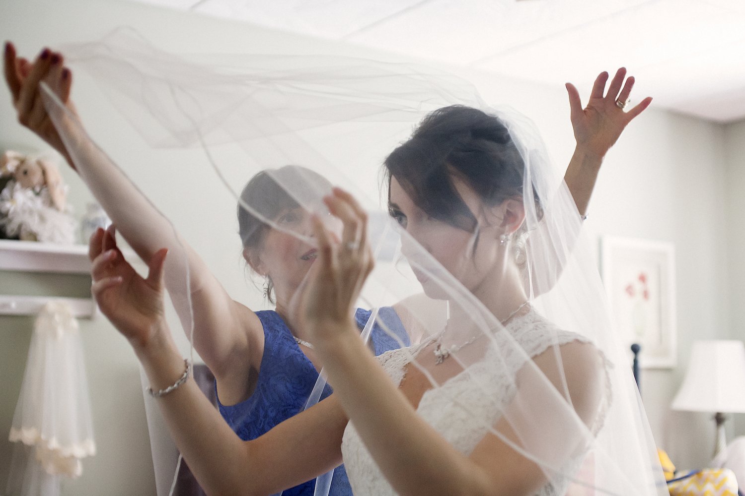 a bride holds her hands up as her mom stands behind her in a blue dress and lifts her long sheer veil over her face before her wedding ceremony.