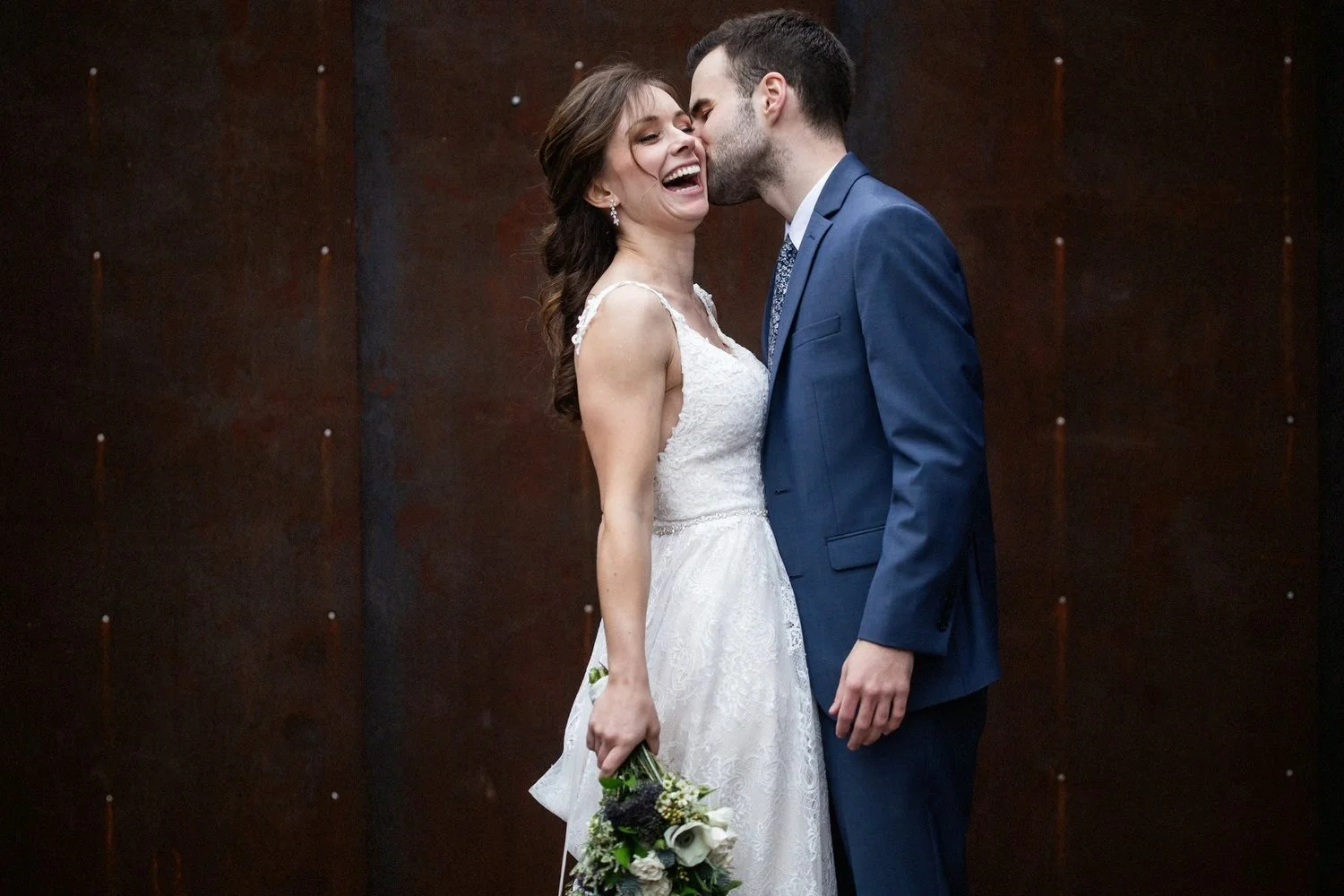 A bride and groom sharing a kiss, with the bride holding a bouquet and smiling as he kisses her cheek, standing against a dark metal background.