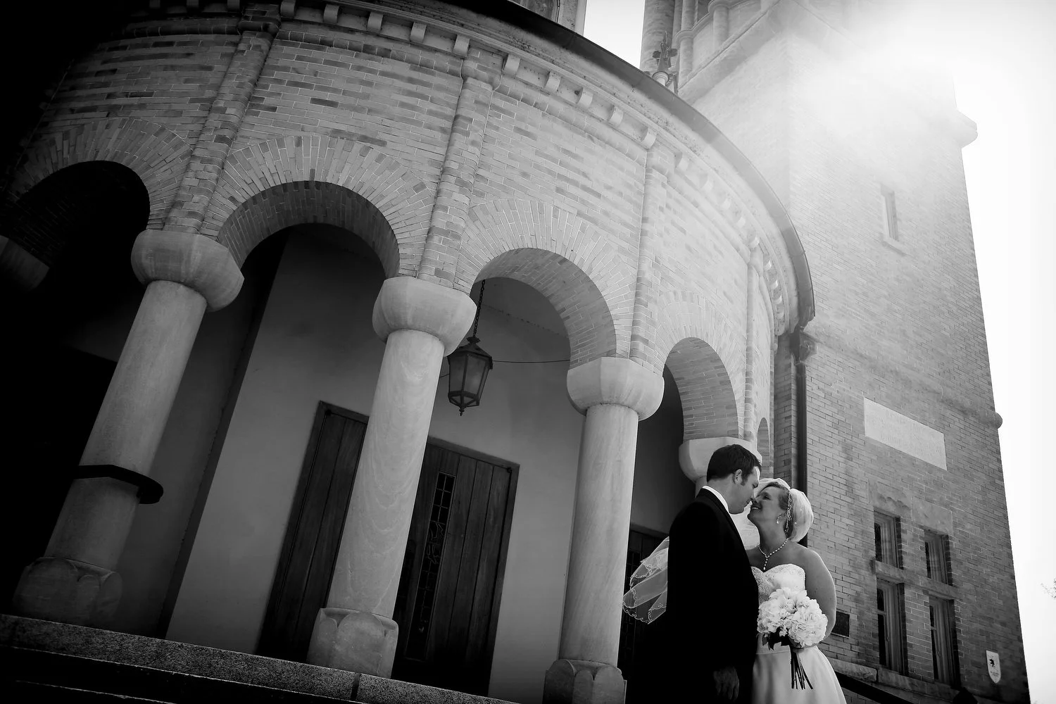 Black and white photo of bride and groom standing close together outside a brick church with ornate columns, sharing a kiss, with sunlight shining behind the building.