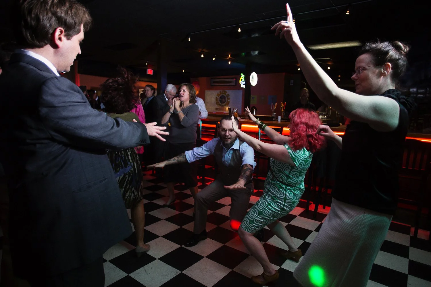 People lean backwards, knees bent and arms outstretched in front of them or in the air as they dance on a black-and-white checkerboard dance floor in a dark room lit with some pink DJ lights in the distance. A woman in a short green dress and reddish