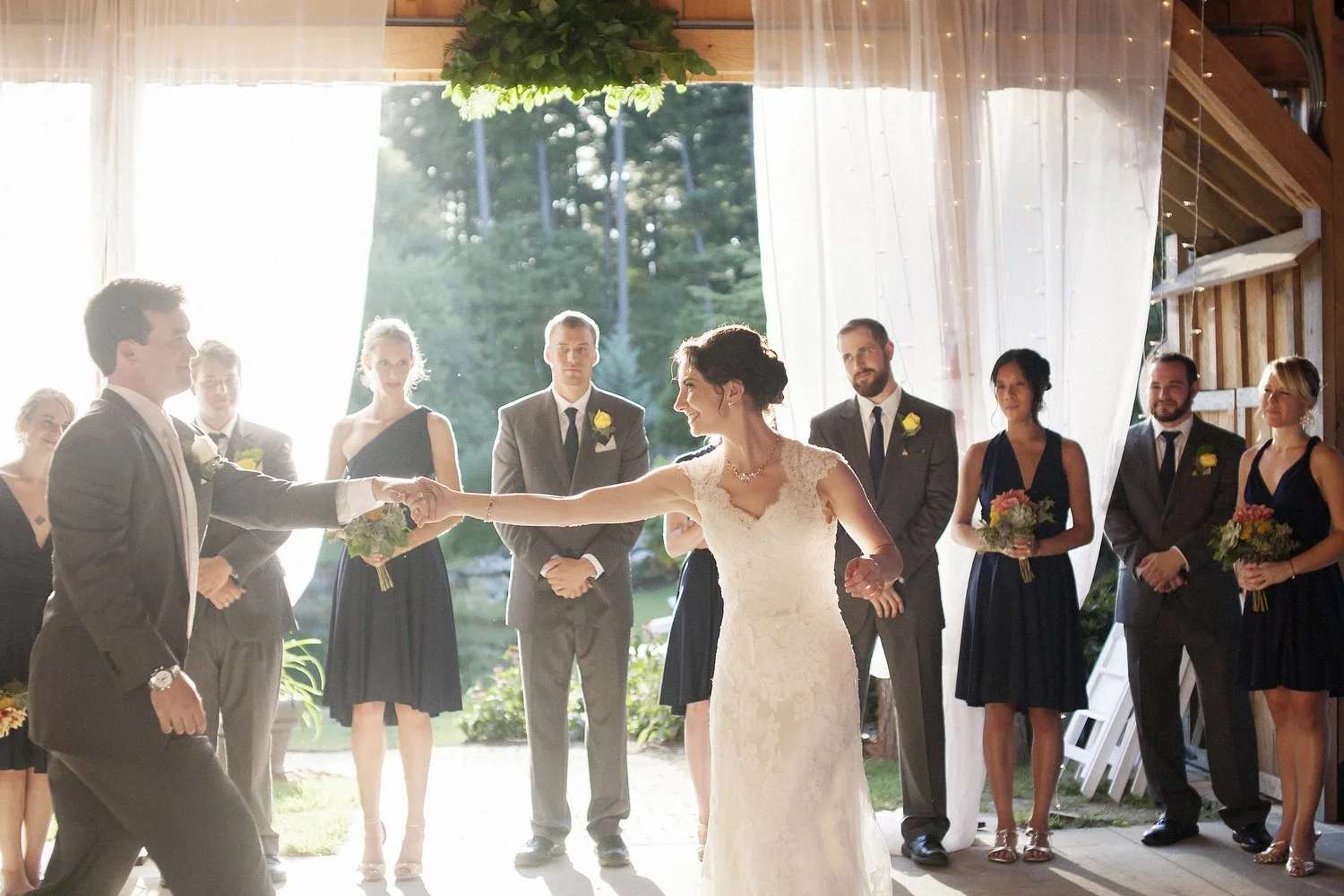Bride and groom dancing during their wedding reception with wedding party and guests watching, set in a rustic venue with open air and natural light. Her arm is outstretched holding his hand.