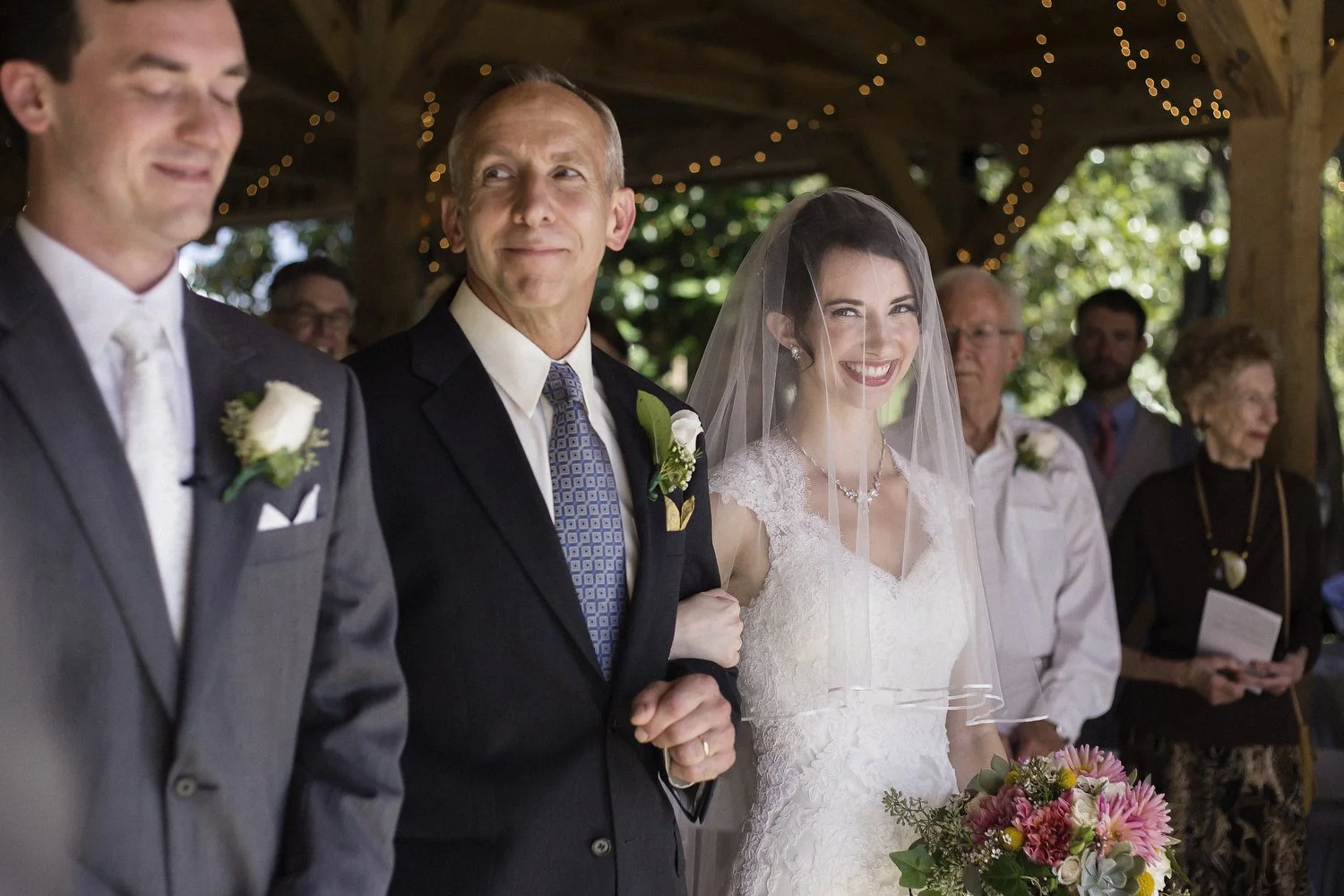 A bride wearing a white lace wedding dress and veil, holds a bouquet of pink and white flowers. She stands next to her father as he looks over at her groom. They are in a wooden outdoor structure decorated with string lights for the wedding ceremony.