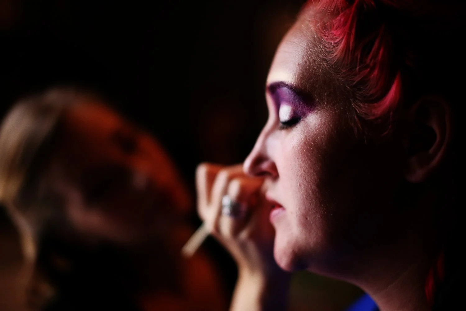 A woman with red hair, closed eyes, and purple eyeshadow has makeup applied with a brush held by a woman in the dark blurred background.