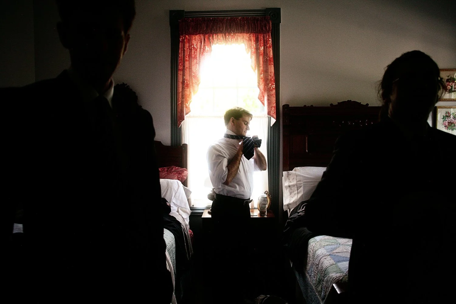 Man adjusting tie in front of window, with two silhouetted people in the foreground, in a bedroom with red lacy curtains and wooden furniture.