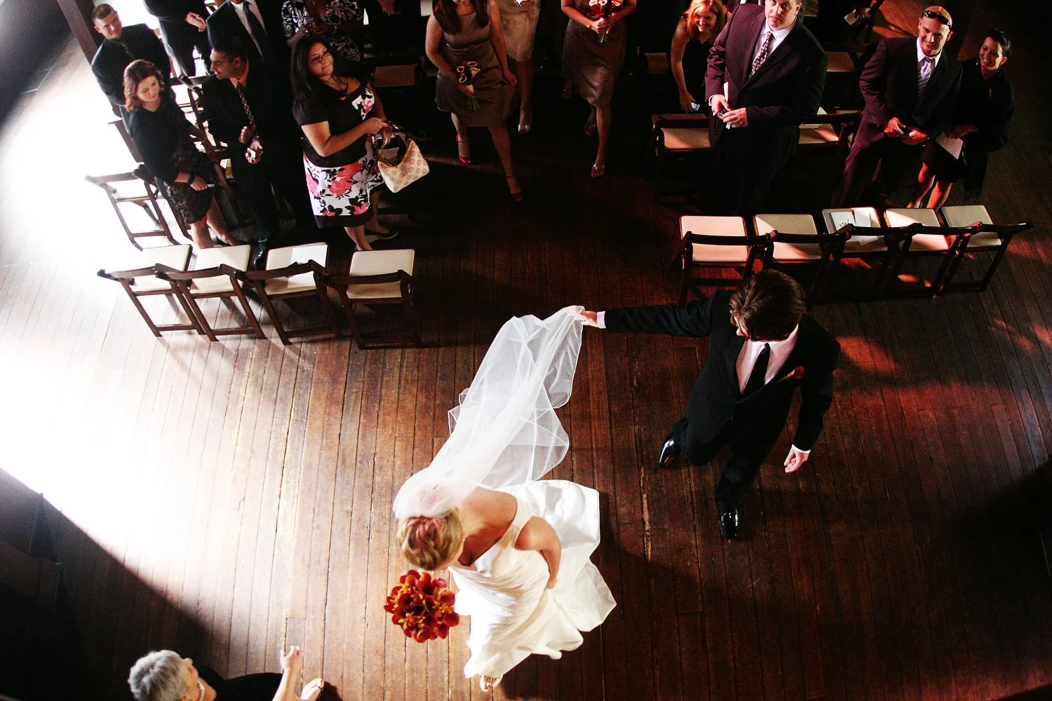 A bride and groom are walking down the aisle at the end of their wedding ceremony. The photo is taken from above and the old dark wooden floors are visible as guests look on from behind them. The bride has a long veil that her groom is holding with a