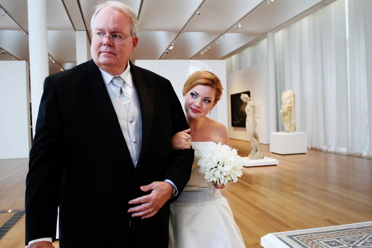 A bride in a white wedding dress holding a bouquet of flowers next to her father in a black suit, inside an art gallery with classical sculptures and white curtains.