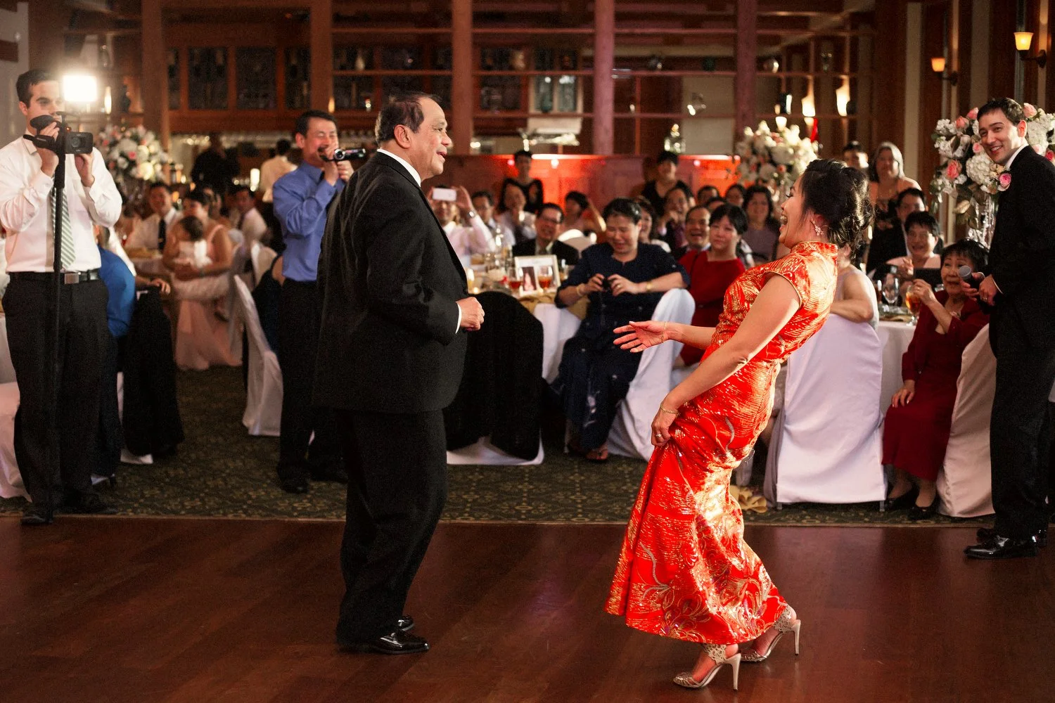 A woman with dark black hair in a fancy updo wearing a long red Asian-styled dress and high heels leans back looking happy as she faces and dances with an older man (maybe her father) in a black suit on a dance floor. There is a flash of light from a