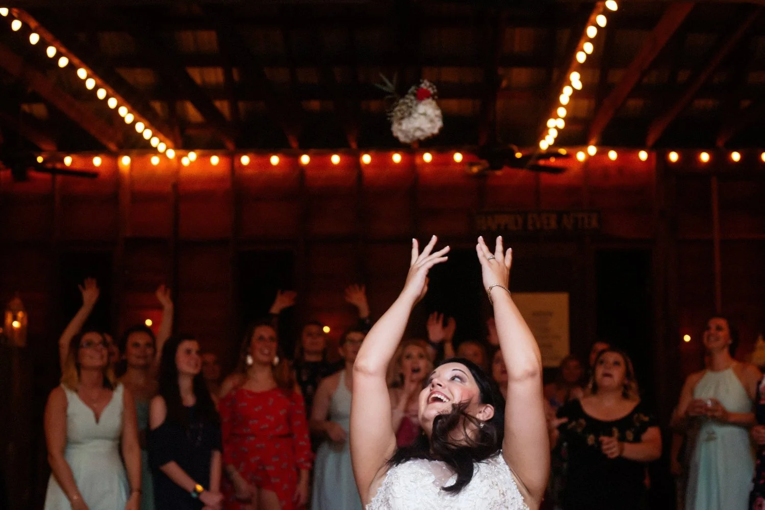 A bride throwing a bouquet during her wedding reception with guests in the background reaching to catch it under warm string lights in a rustic barn setting.