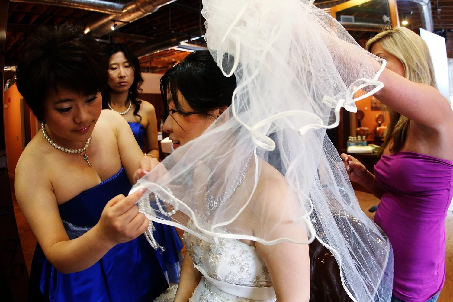 Woman in a white wedding dress getting ready, surrounded by women in colorful dresses helping her adjust her tulle veil and jewelry in a warmly lit room.