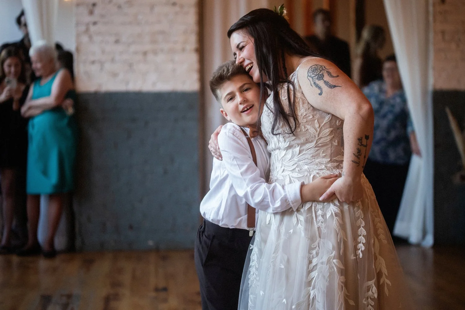 A woman and her young son sharing a hug at a celebration or wedding reception, smiling happily, with guests in the background. She is wearing a lacy wedding dress, and he has dress pants and a dress shirt.